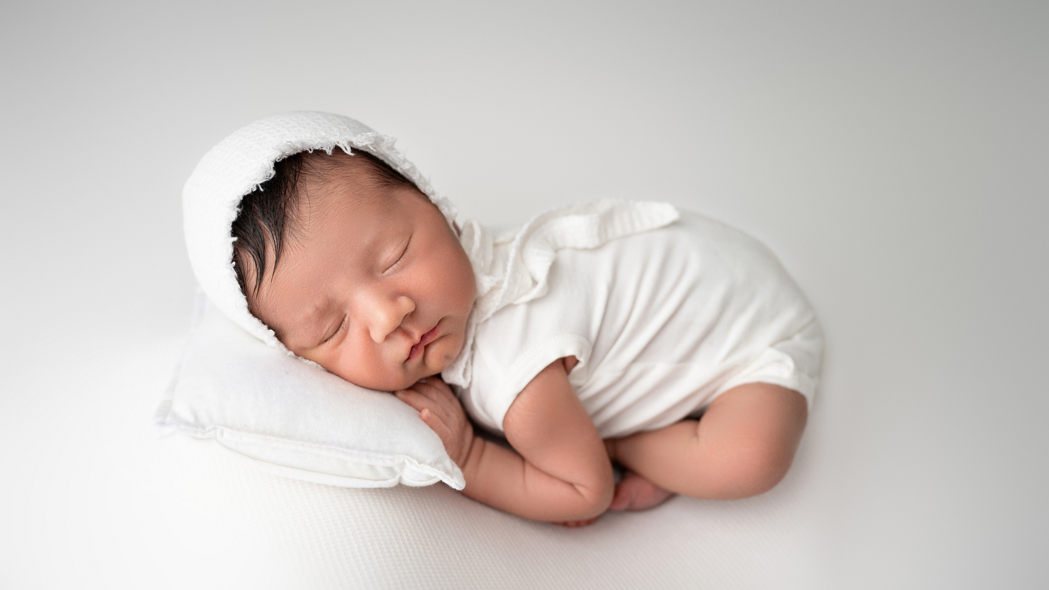 Sleeping newborn in white outfit resting on soft pillow in minimalist setting.