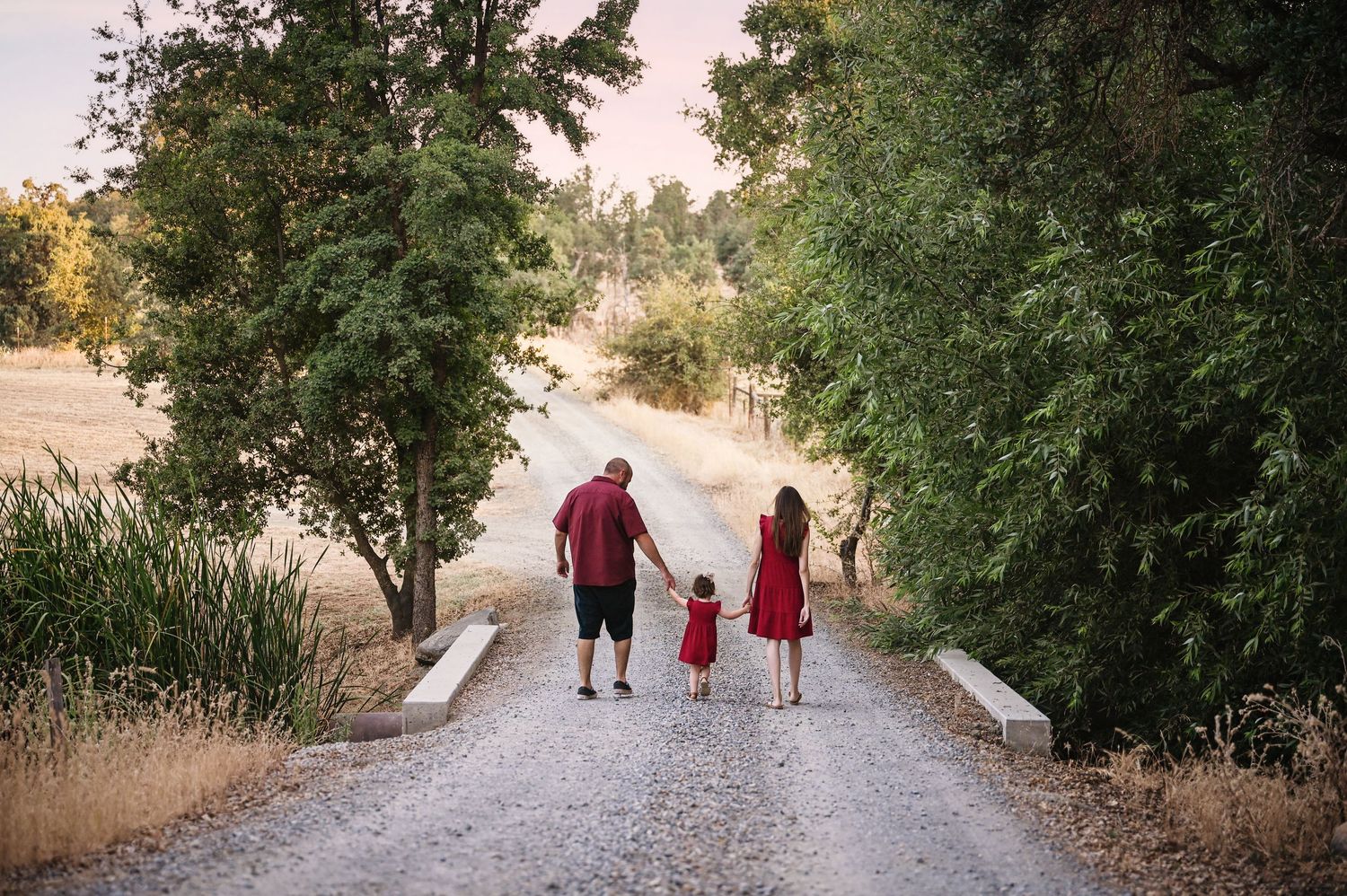 Three people in red walk on a tree-lined path together during sunset. Visalia Family Photographer