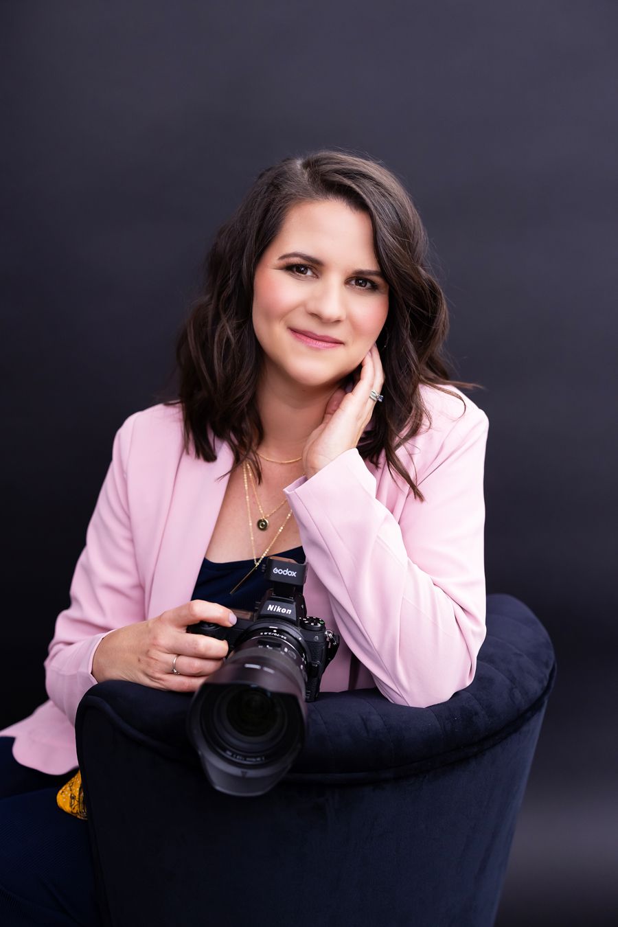 Professional maternity & newborn photographer in Lemoore, CA in pink blazer poses with camera against dark background in studio portraits.