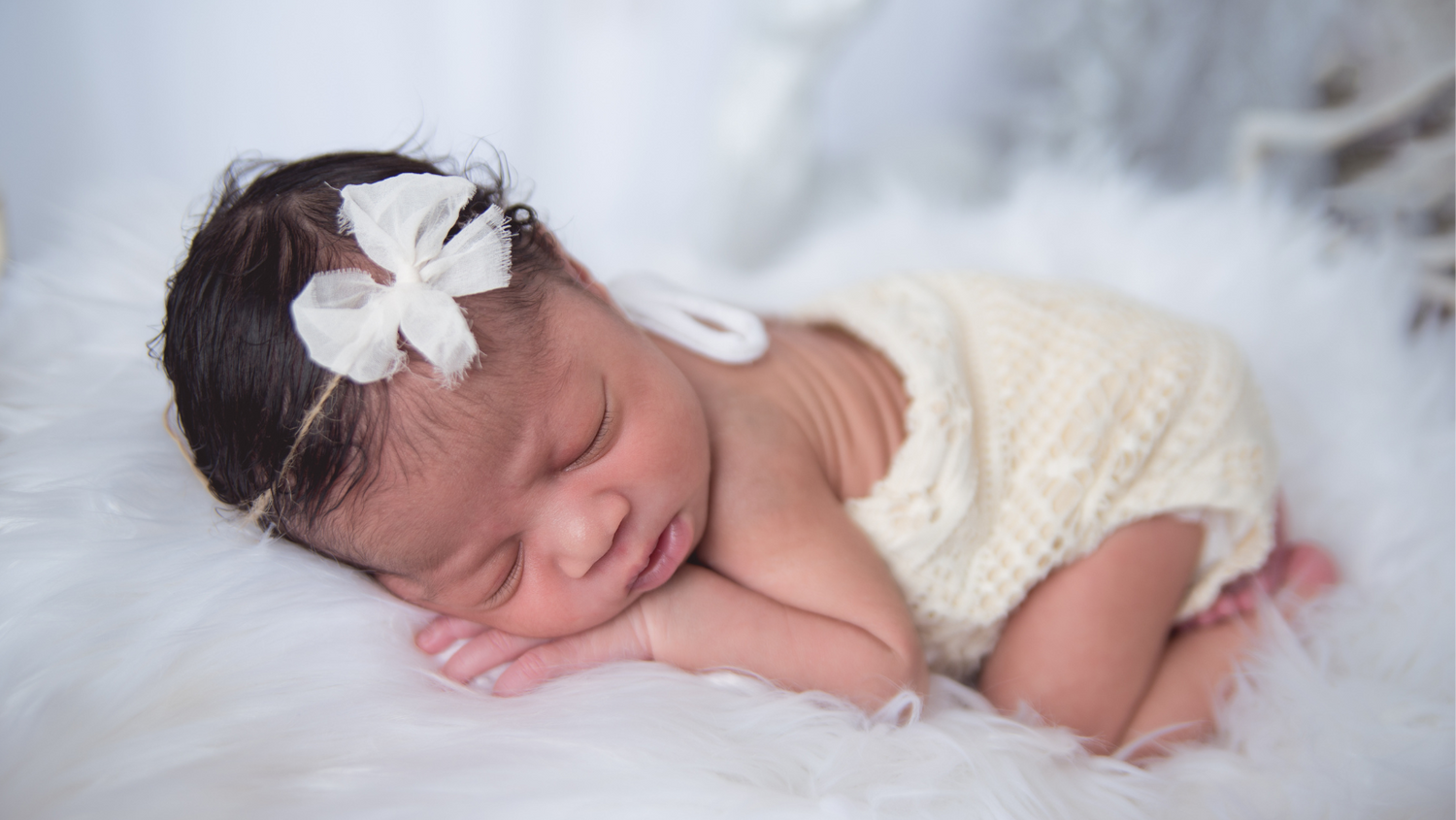 Sleeping newborn in cream-colored outfit with butterfly hair accessory on white fabric.