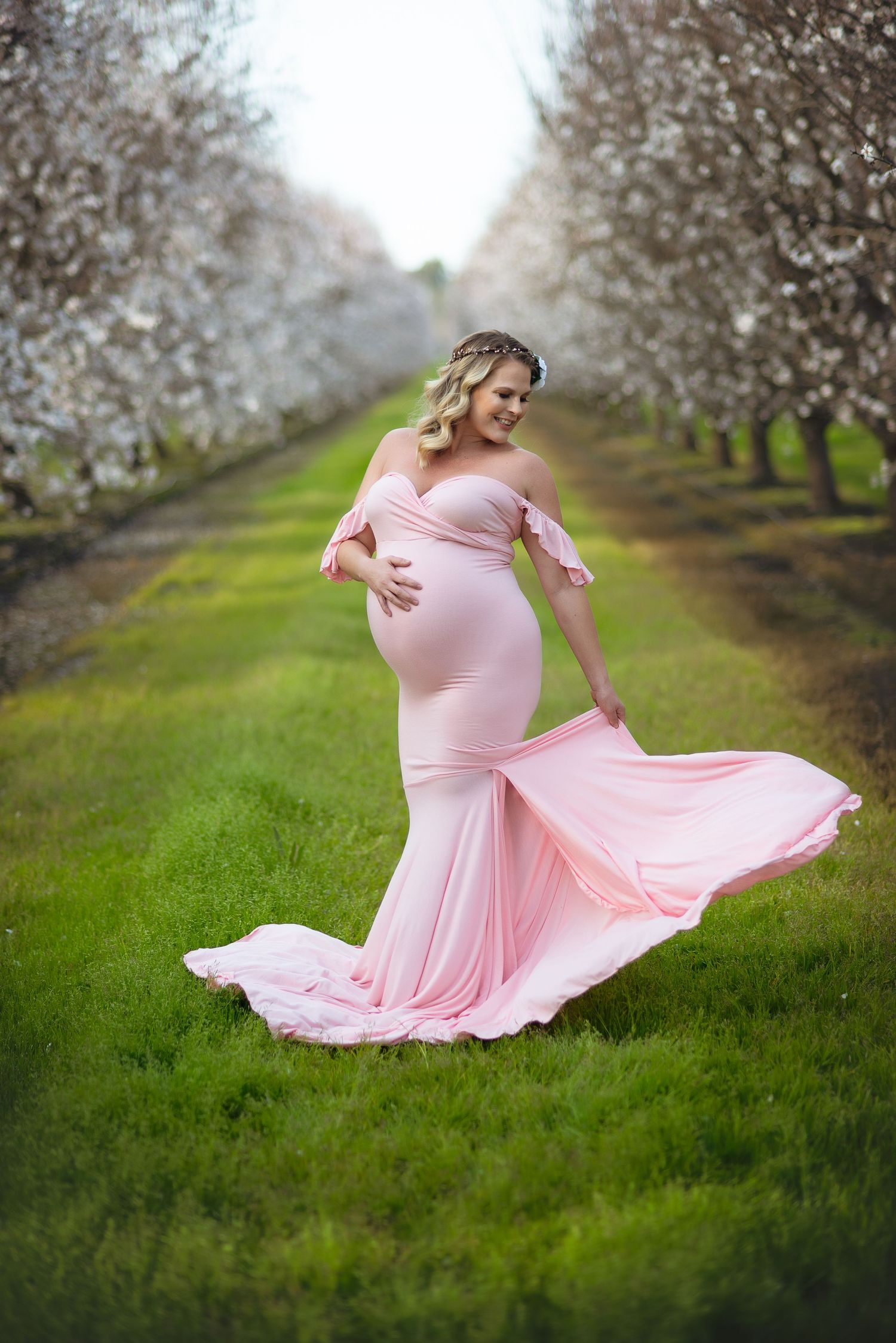 A flowing pink maternity dress twirls gracefully among blooming almond trees in a spring orchard in Lemoore, CA.