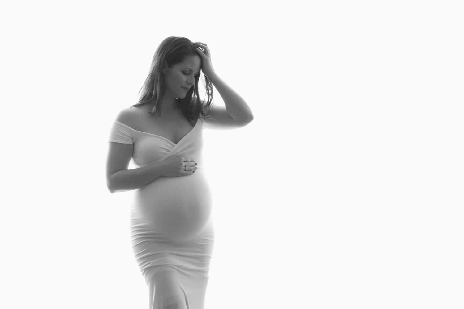 Artistic black and white maternity photo of an expecting mother in a form-fitting white dress against a bright background. Fresno Maternity