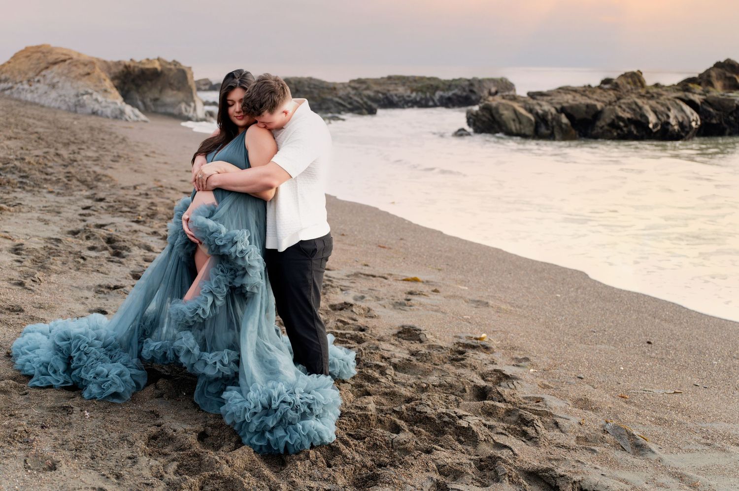 Couple sharing intimate moment on beach at sunset with flowing teal dress.
