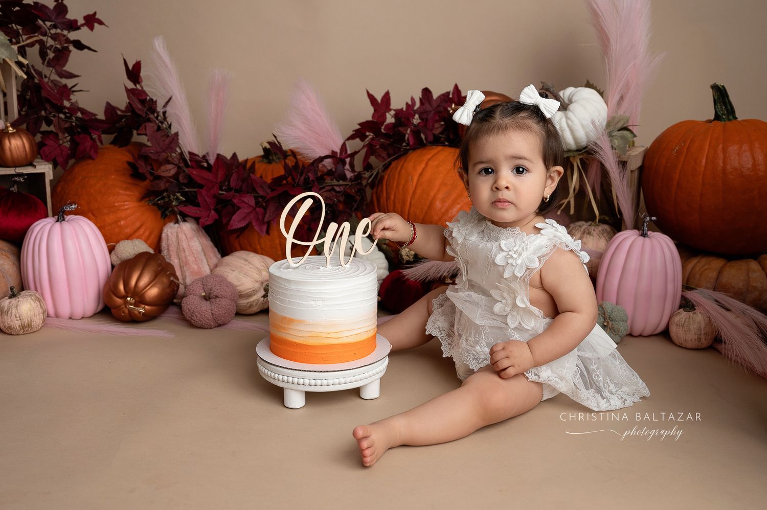 Fall themed cake smash photography setup with white cake, orange and pink pumpkins, and burgundy foliage decorations. Hanford Baby Photos