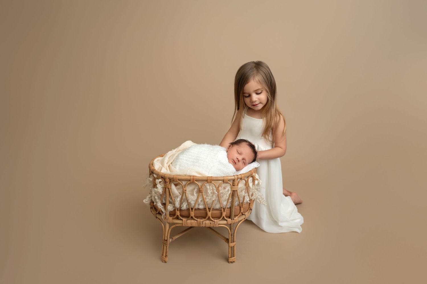 Young child in white dress looking at sleeping newborn in wicker bassinet.