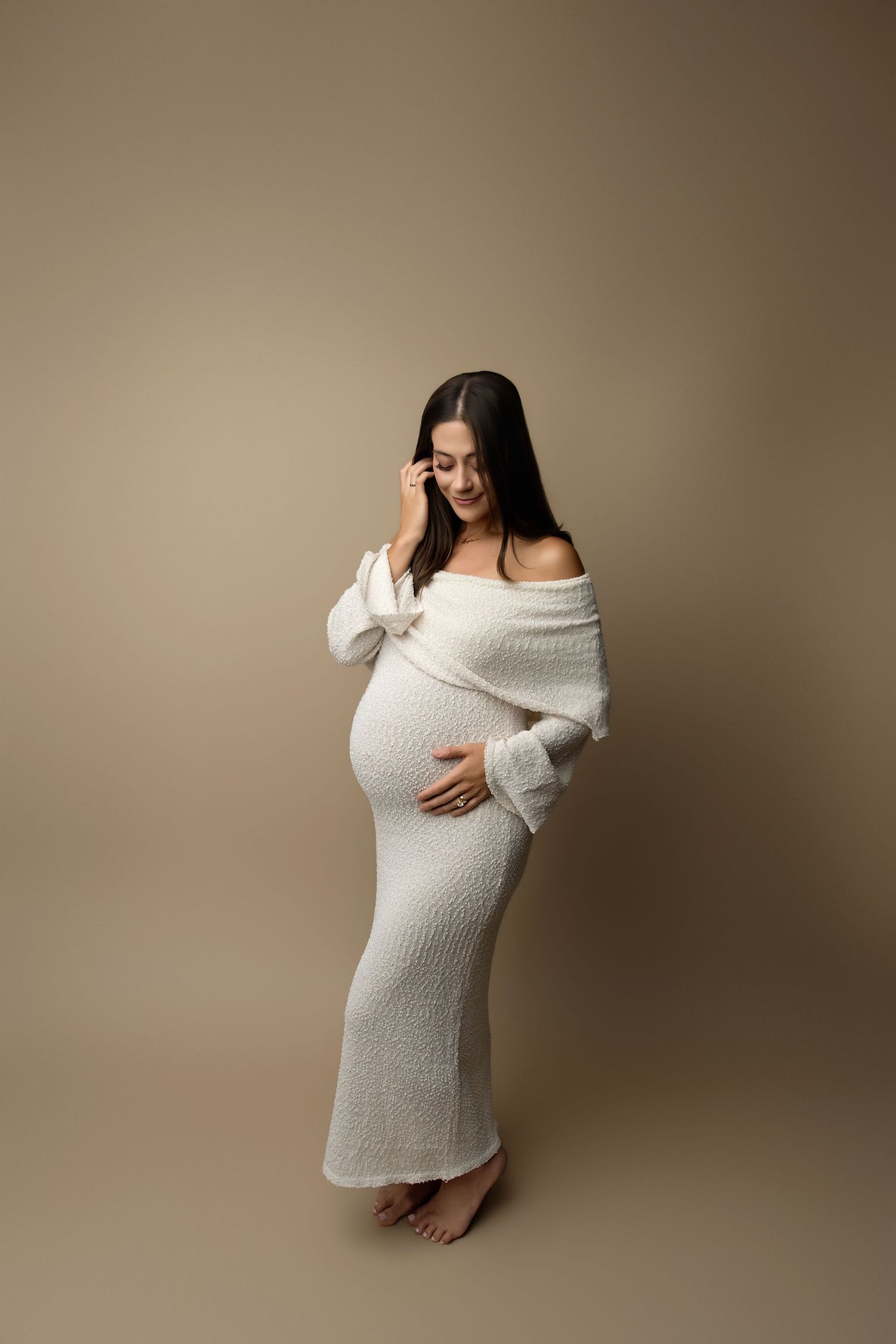 Pregnant woman in white off-shoulder knit dress cradling baby bump, posing barefoot against beige background.