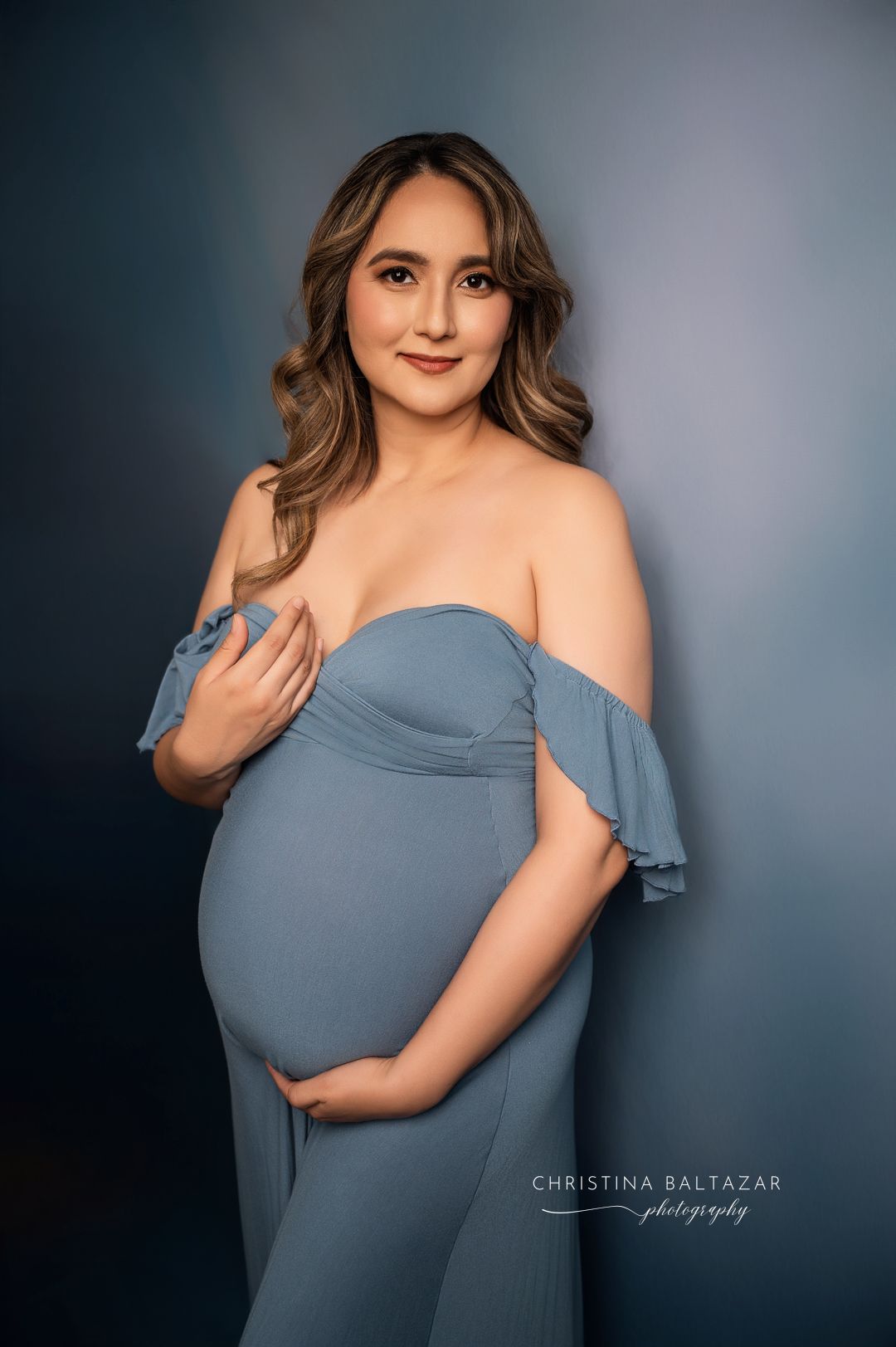 Elegant maternity portrait featuring an off-shoulder dusty blue gown against a dark background. Hanford Maternity Photographer