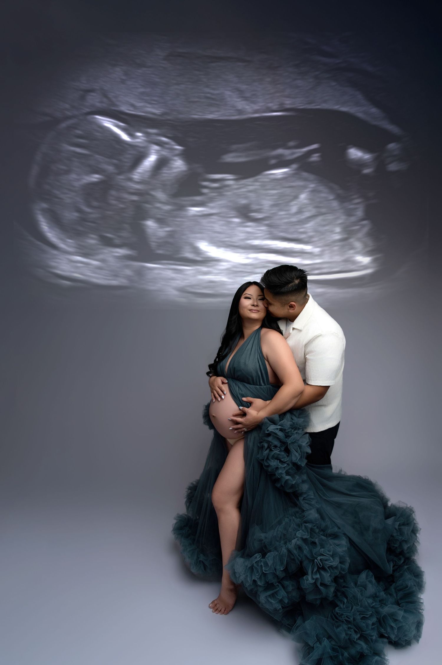 Pregnant couple embracing in a studio, woman in teal tulle gown, ultrasound image projected above them.