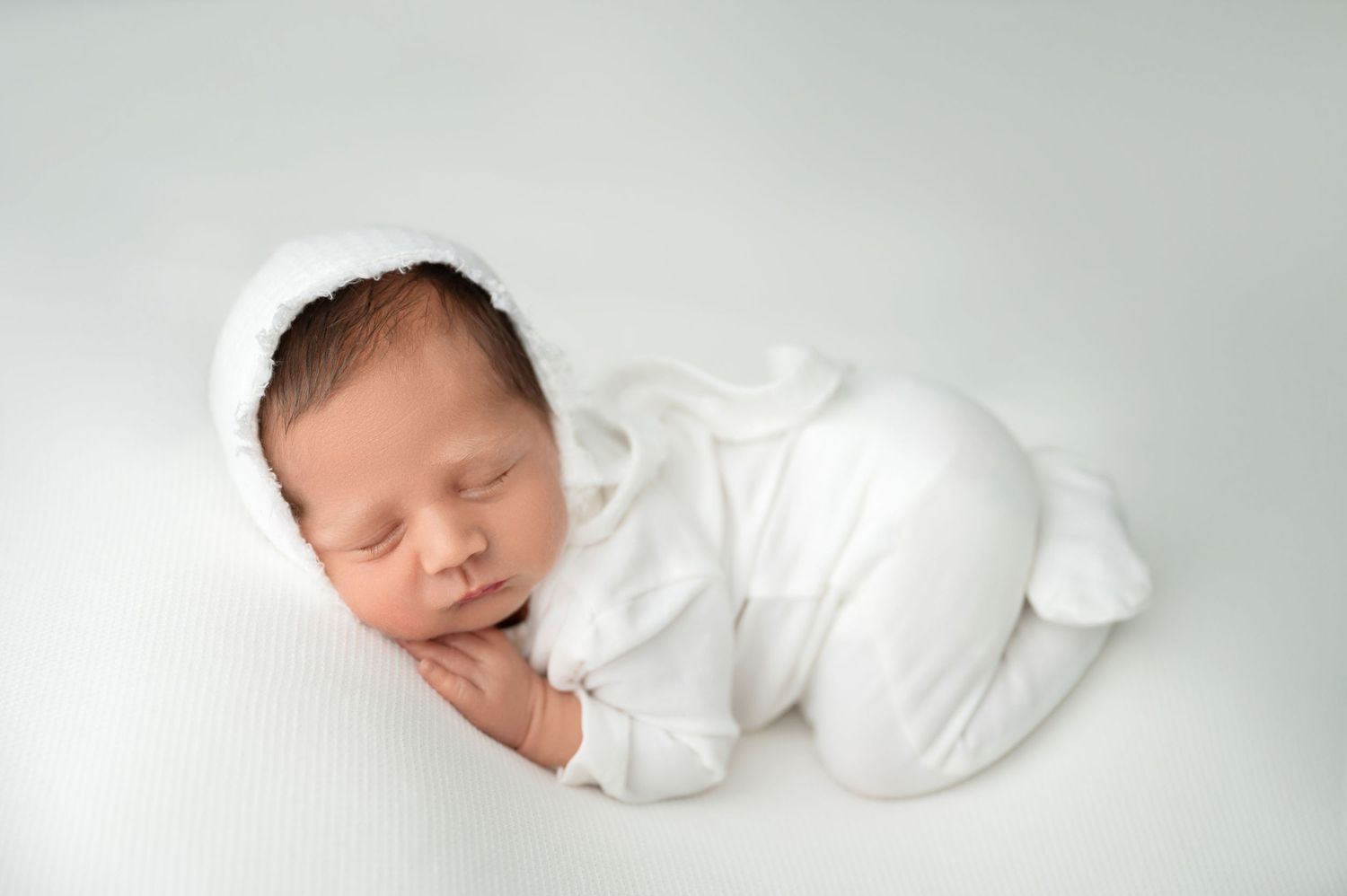 Sleeping baby in white outfit curled up on light studio backdrop.