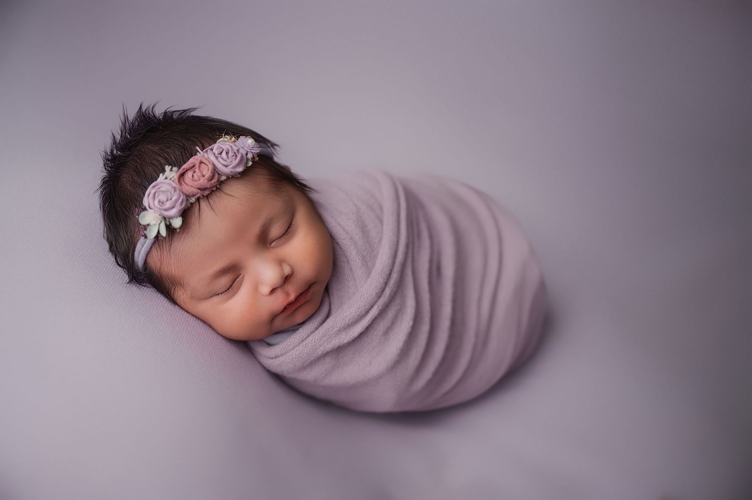 Sleeping newborn wrapped in soft purple swaddle with floral headband on purple backdrop.