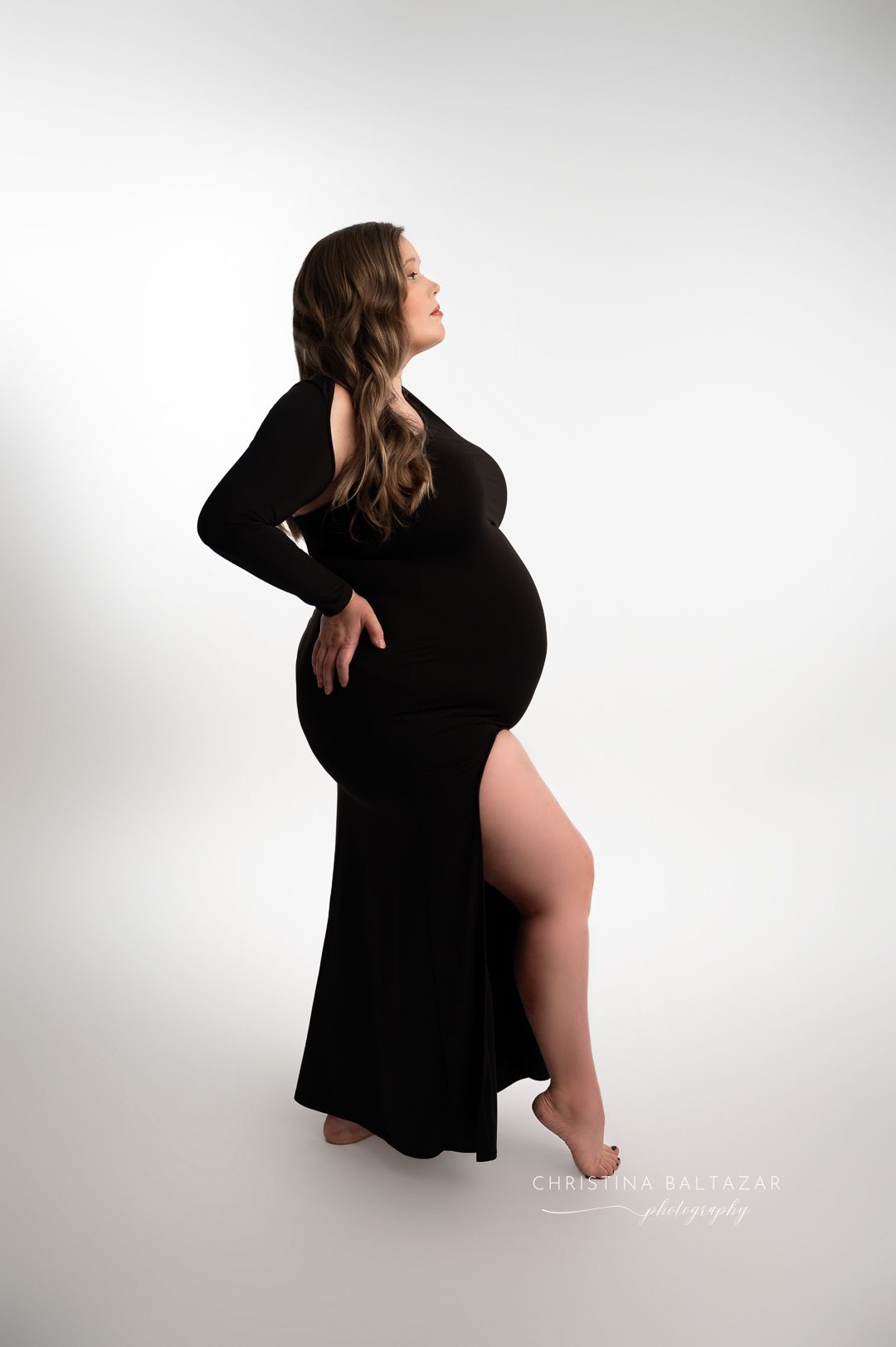 Elegant maternity photo of a woman in a flowing black dress posing against a white background in a studio setting. Fresno Maternity Photos