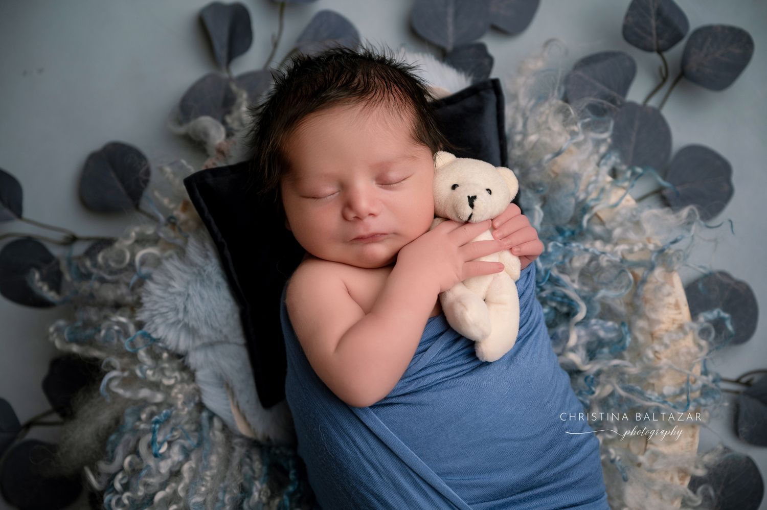 A newborn baby peacefully sleeps while holding a small teddy bear, wrapped in blue fabric. Fresno Newborn Photographer