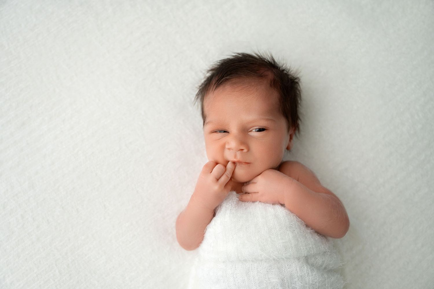 Newborn baby swaddled in white with hand near mouth on light background.