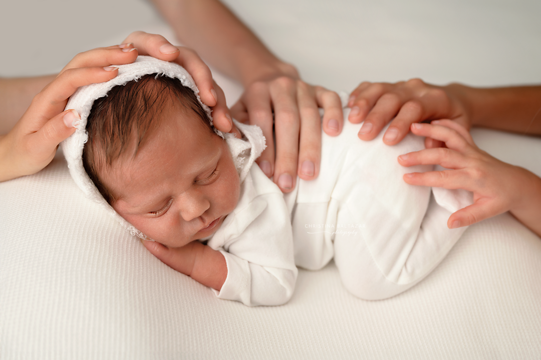 Sleeping newborn in white outfit being gently touched by caring hands.