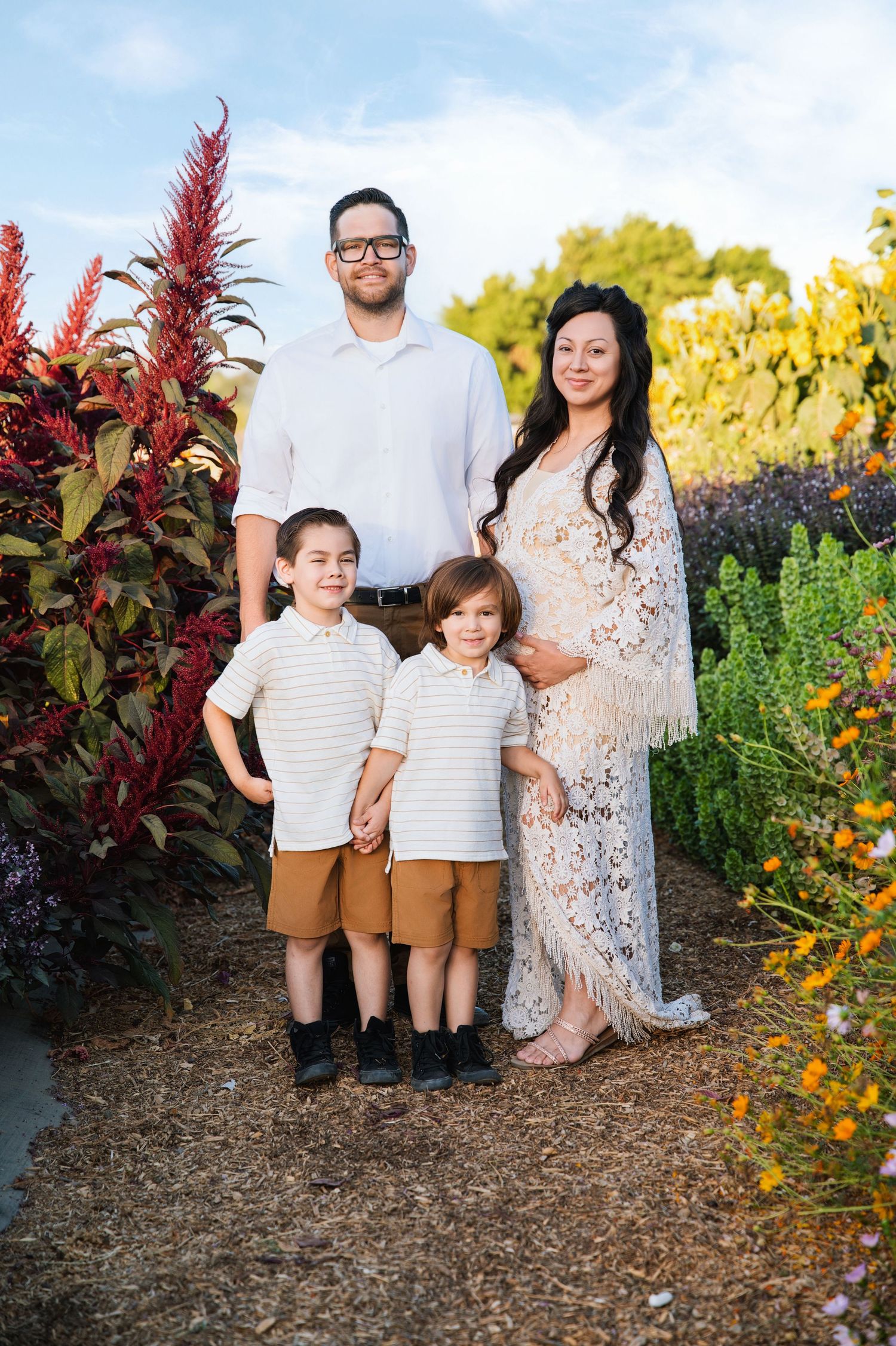 Family poses together in a garden with red flowers and yellow blooms during a sunny outdoor portrait session. Hanford Family Photographer