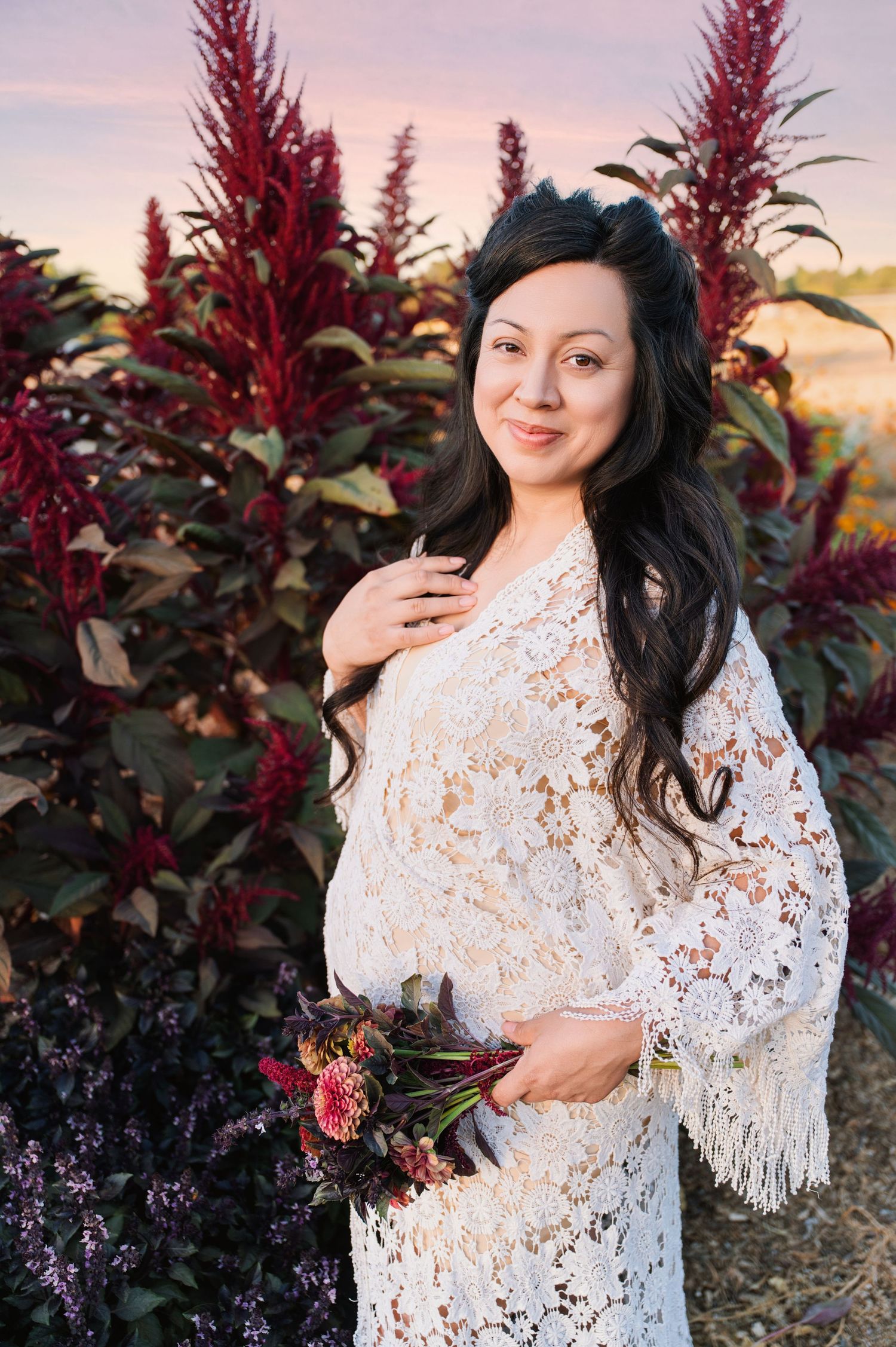 A woman in a white lace dress poses among vibrant red foliage during a sunset photoshoot.