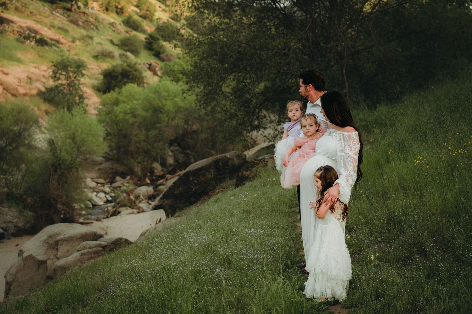 White-dressed figures stand near a rocky stream bed on a grassy hillside. Fresno Family Photography