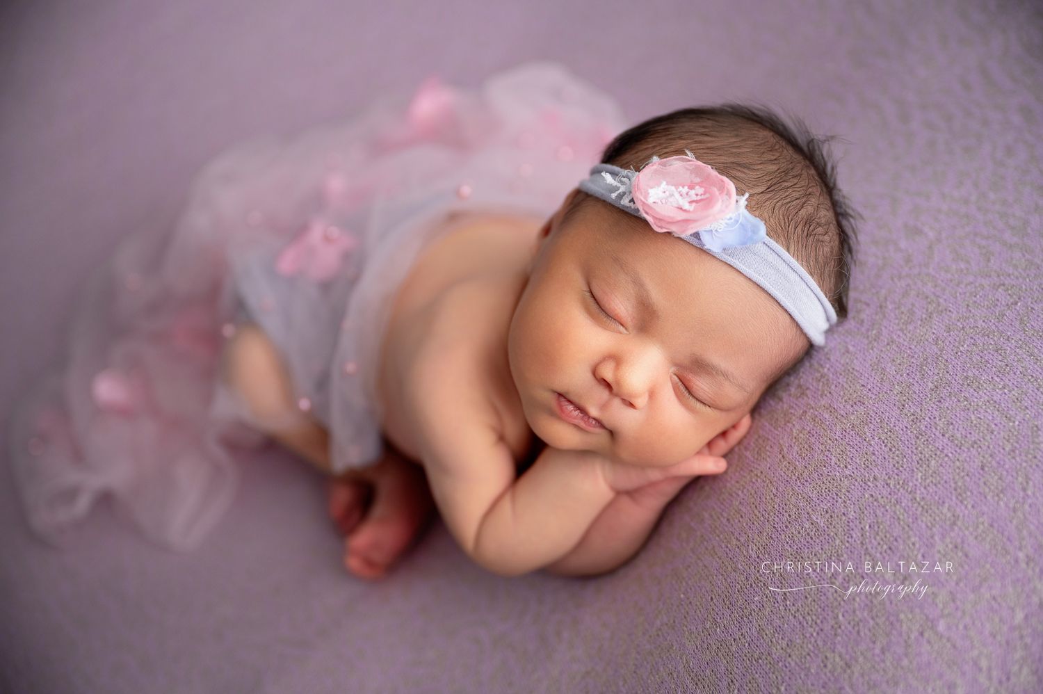 A sleeping newborn wearing a delicate tulle skirt and blue headband rests peacefully on a soft purple blanket. Fresno Newborn Photographer