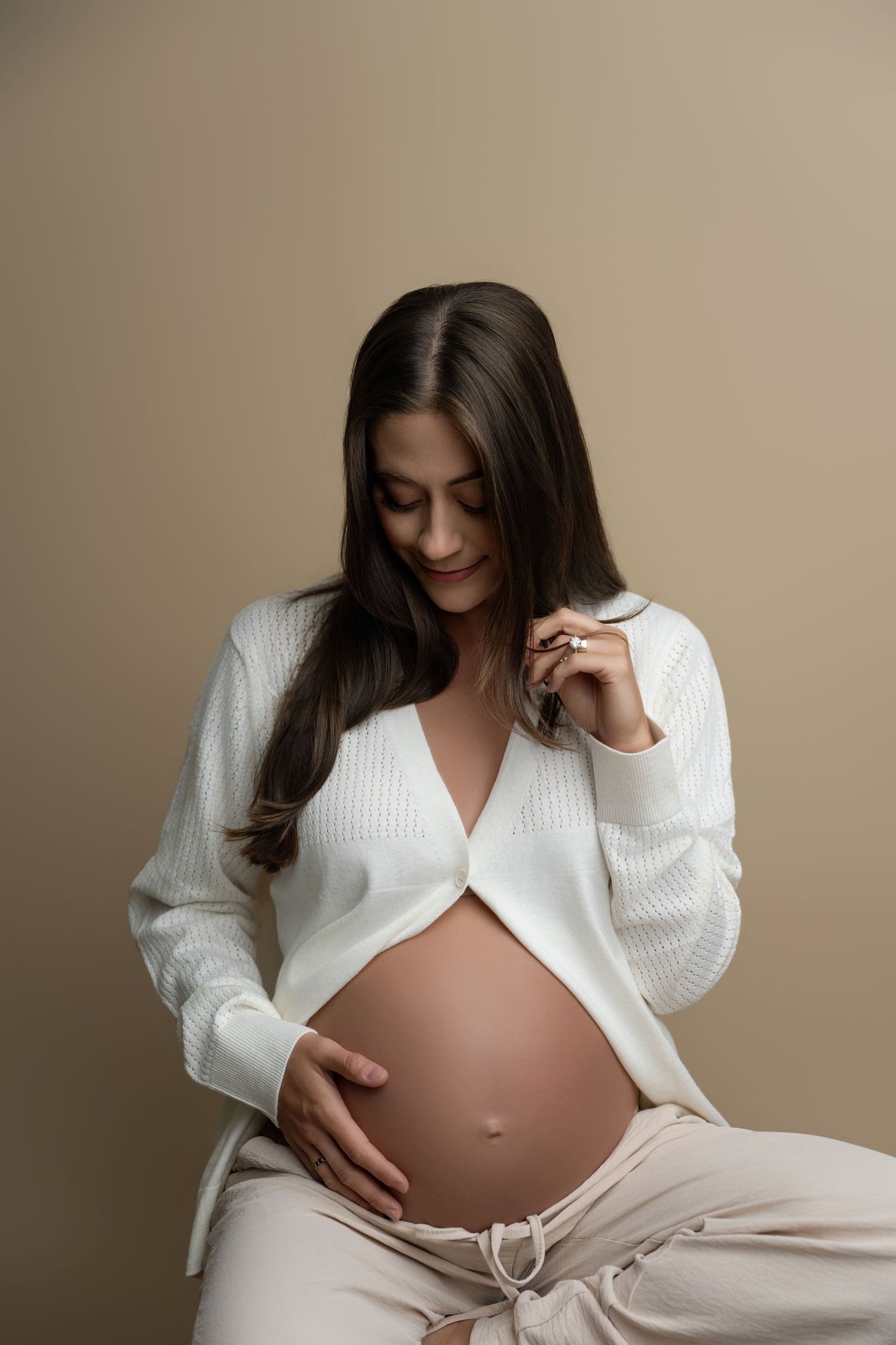 Pregnant woman in white cardigan cradling her baby bump, smiling softly against a neutral beige background.
