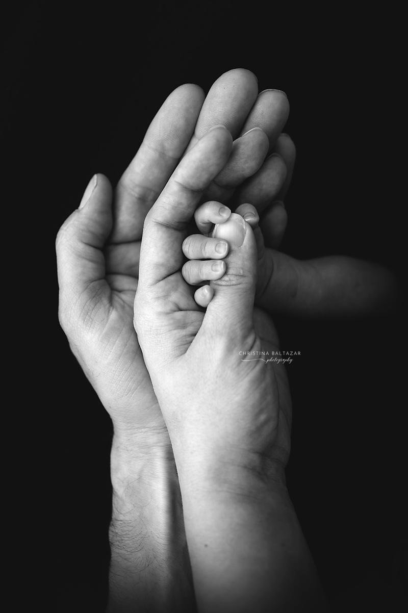 Black and white close-up of adult and baby hands tenderly touching against dark background.