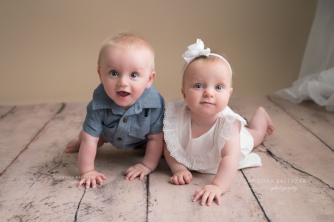 Two babies in dressy outfits sit together on a wooden floor with one wearing denim and the other in white. Fresno Baby Photographer