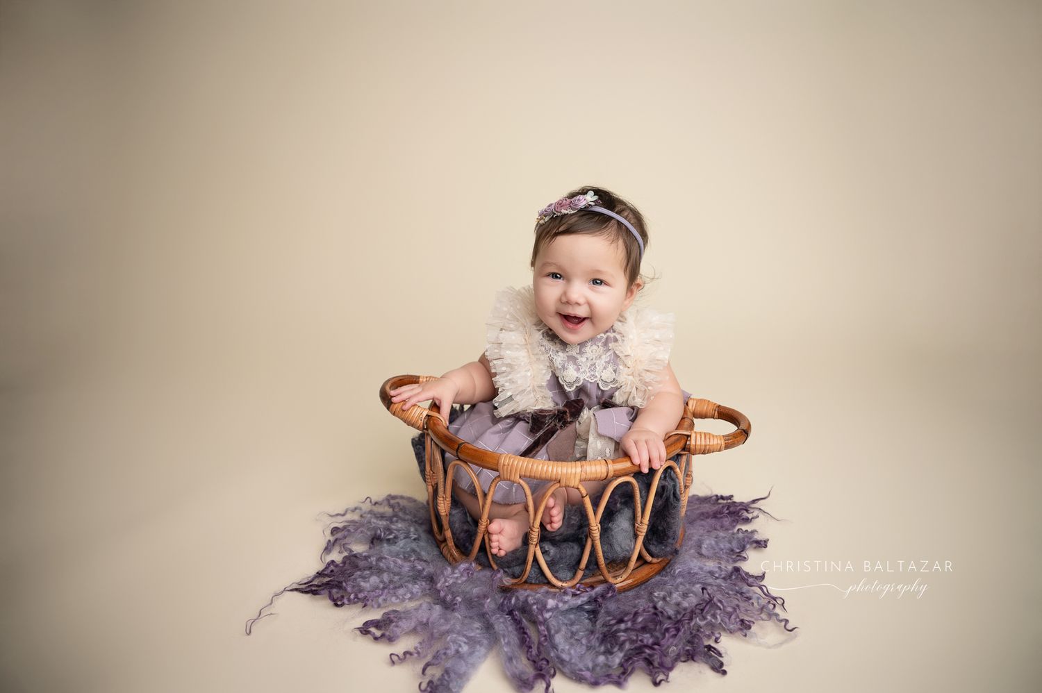 Smiling baby sitting in brown wicker basket wearing white tulle outfit with purple feathers spread underneath on beige backdrop. Fresno baby
