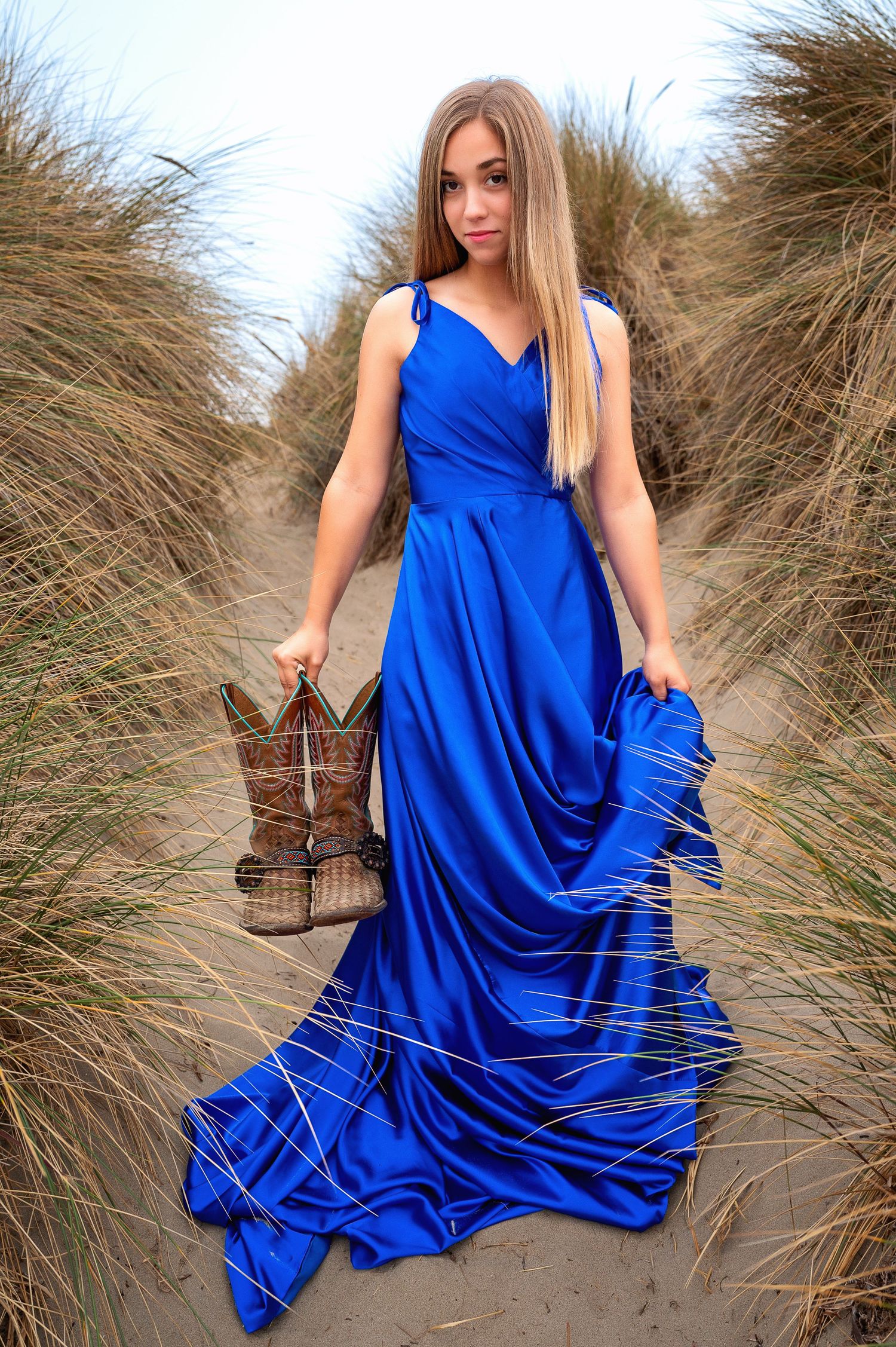 Long blue dress flows dramatically among beach grass while holding vintage boots.