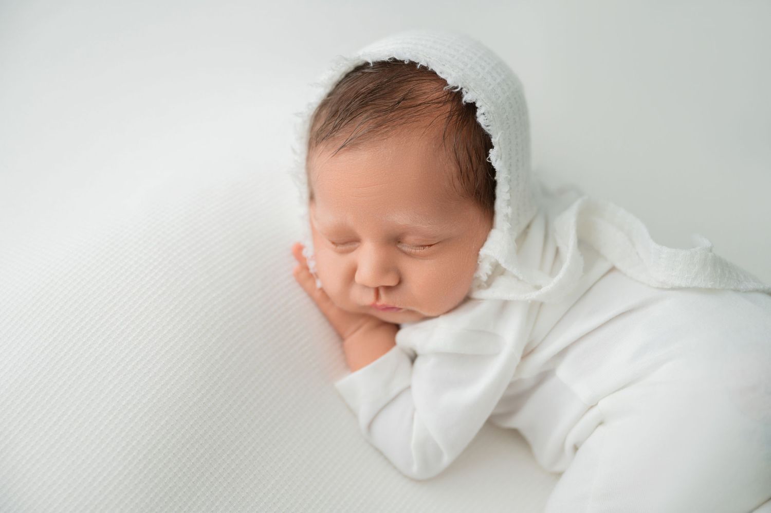Sleeping newborn in white outfit resting head on hand during photo session.