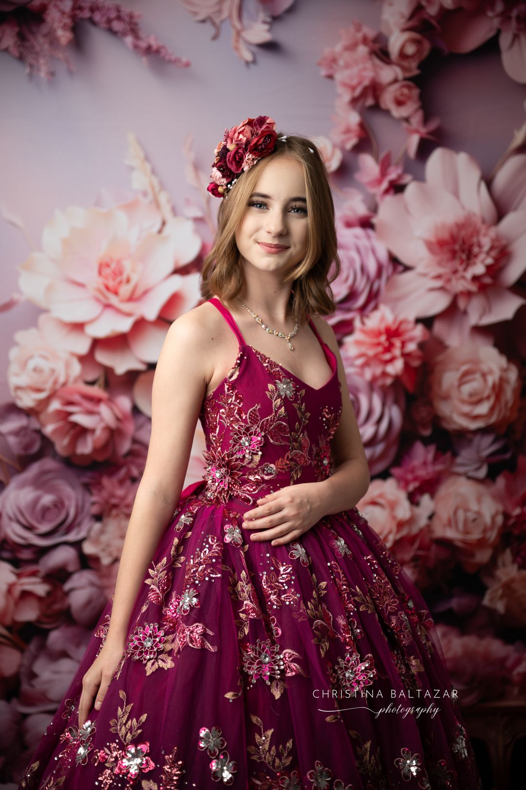 Elegant burgundy ball gown with gold embroidery photographed against romantic pink floral backdrop.