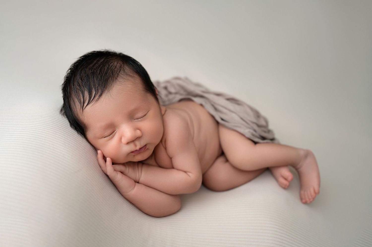 A sleeping newborn baby lies peacefully on their side against a soft cream backdrop with a delicate light wrap. Fresno Newborn Photography