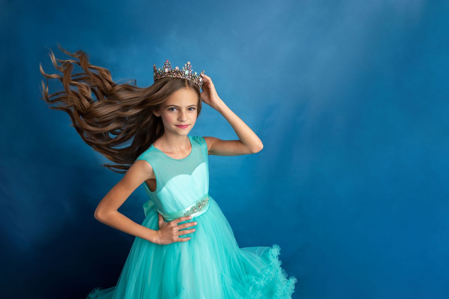 Elegant figure in turquoise ball gown with flowing hair poses against blue backdrop.