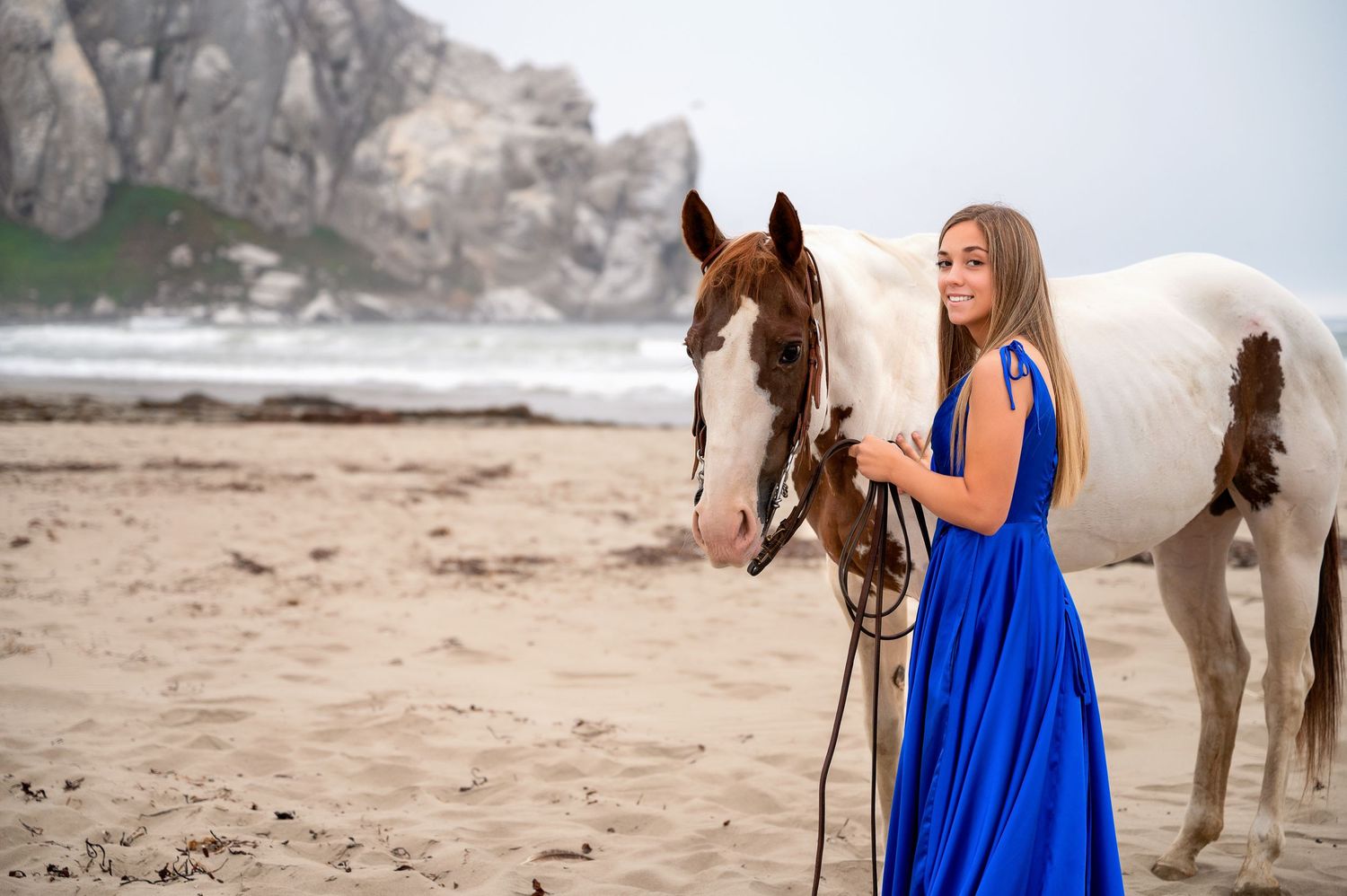 A person in a flowing blue dress stands with a paint horse on a misty beach with rocky cliffs in the background.