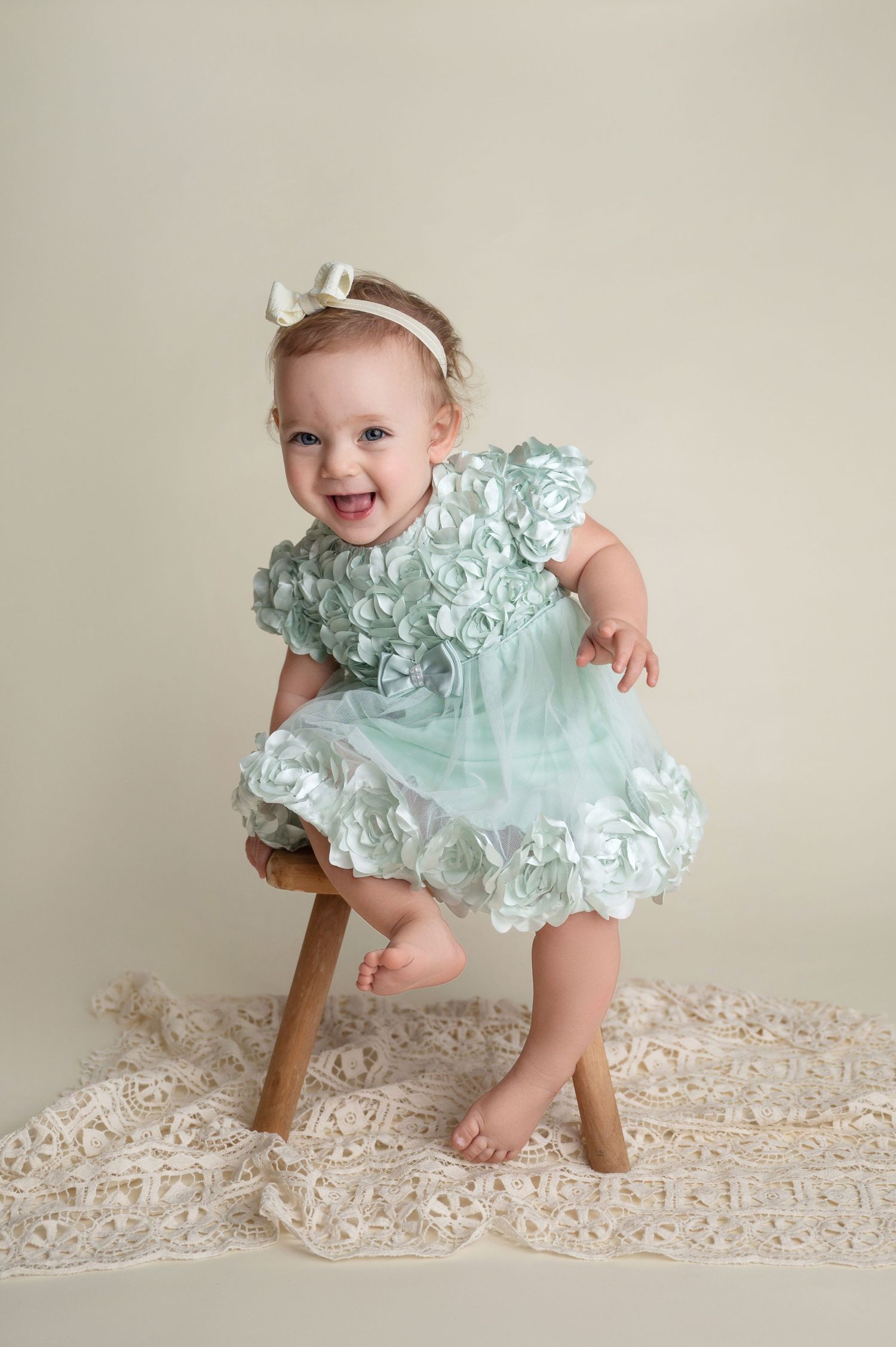 Baby in mint green ruffled dress poses on wooden stool against neutral backdrop with lace details.