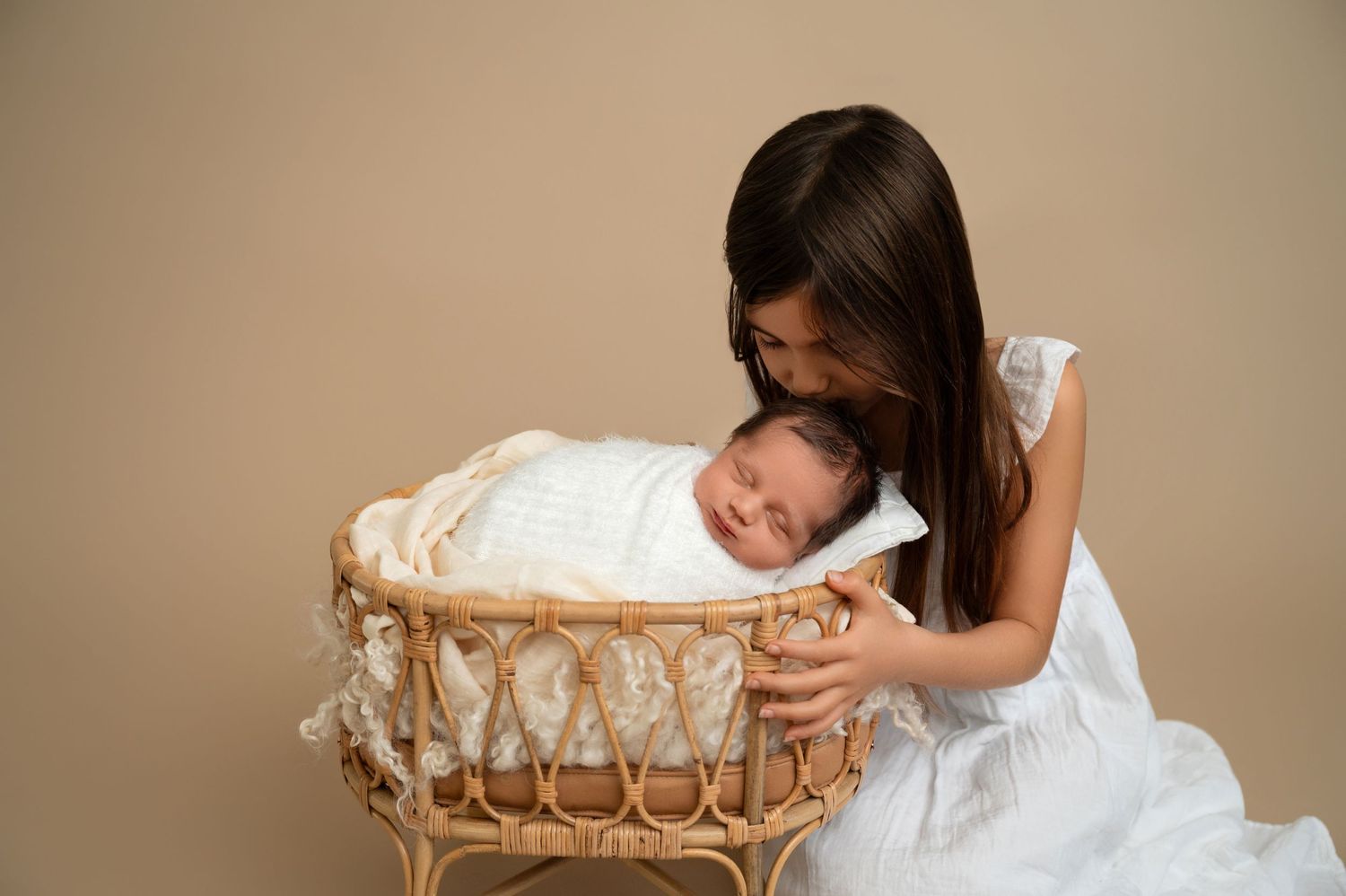 A tender moment as a girl in white dress leans over a wicker bassinet holding a sleeping baby.