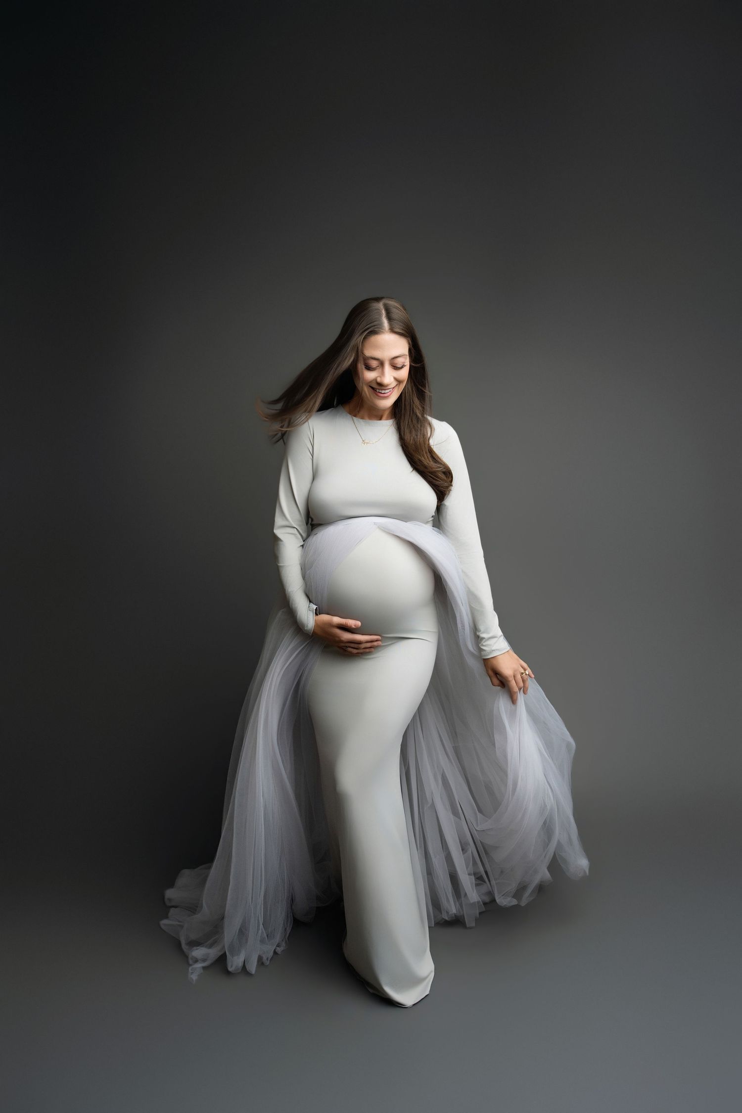 Smiling pregnant woman in elegant white gown with flowing tulle, cradling baby bump in professional studio maternity photo in Hanford, CA.
