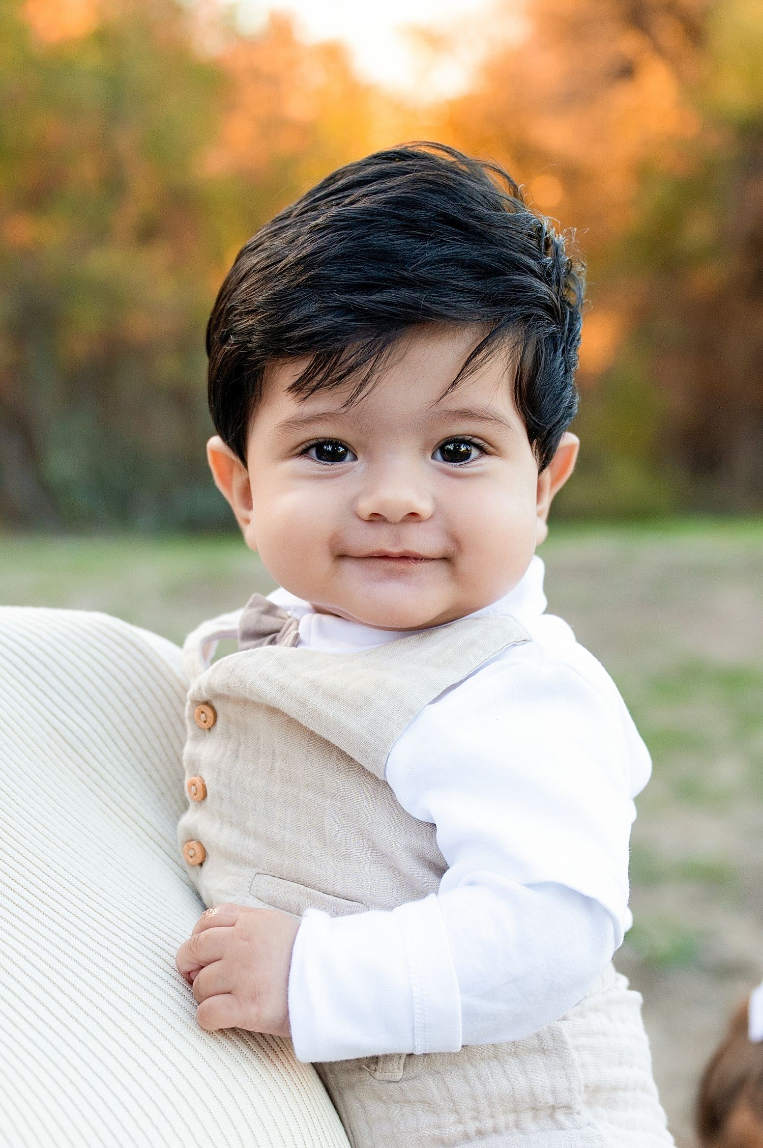 A baby in a cream-colored sweater and white shirt sits outdoors during golden hour with autumn foliage in the background. Fresno Family Phot