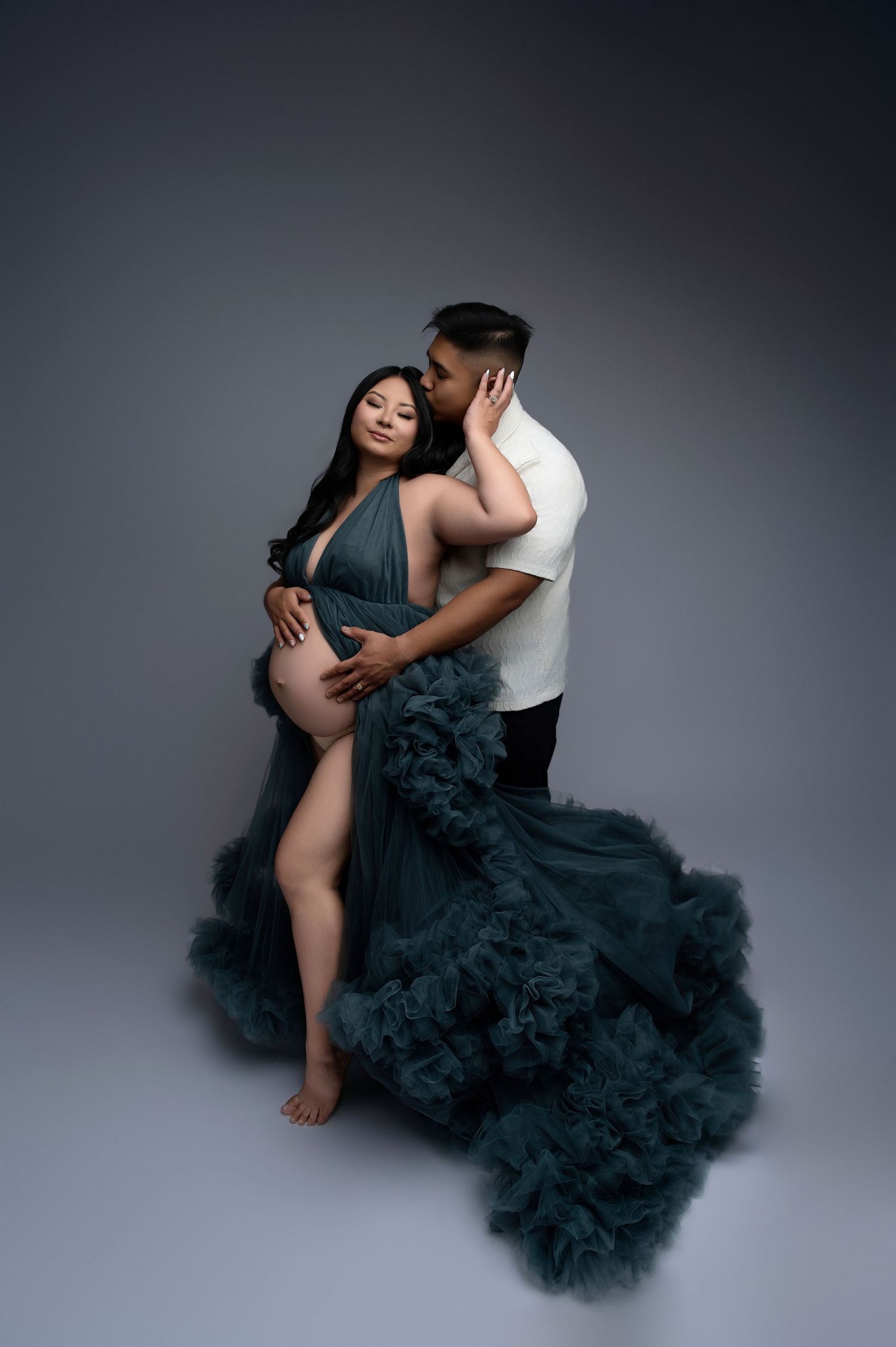 Couple in maternity photo shoot, woman in teal ruffled gown cradling baby bump while partner kisses her cheek in studio.