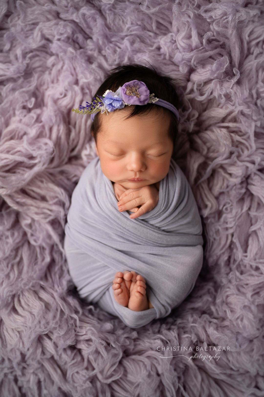 A sleeping newborn wrapped in soft gray fabric lies on a textured lavender background wearing a delicate floral headband. Fresno Newborn Pho
