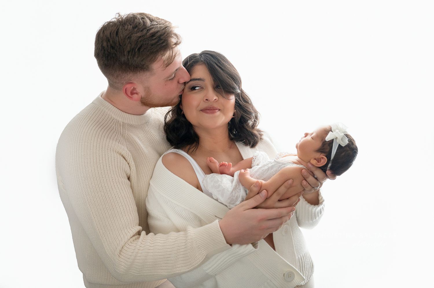 Affectionate couple in cream colored outfits holding a newborn baby against white background. Hanford Newborn Photographer
