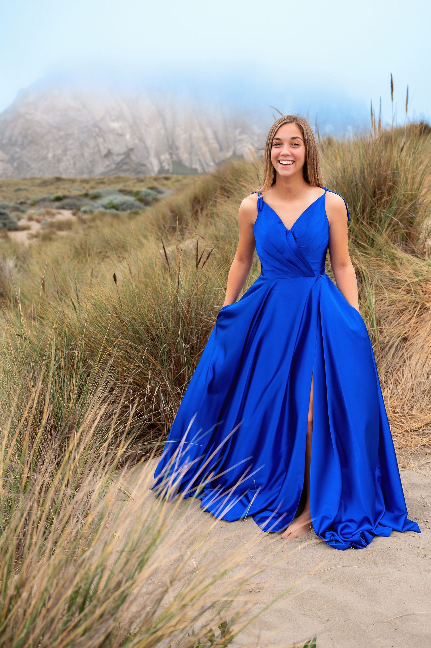 Royal blue gown billows in the wind against coastal mountain landscape.