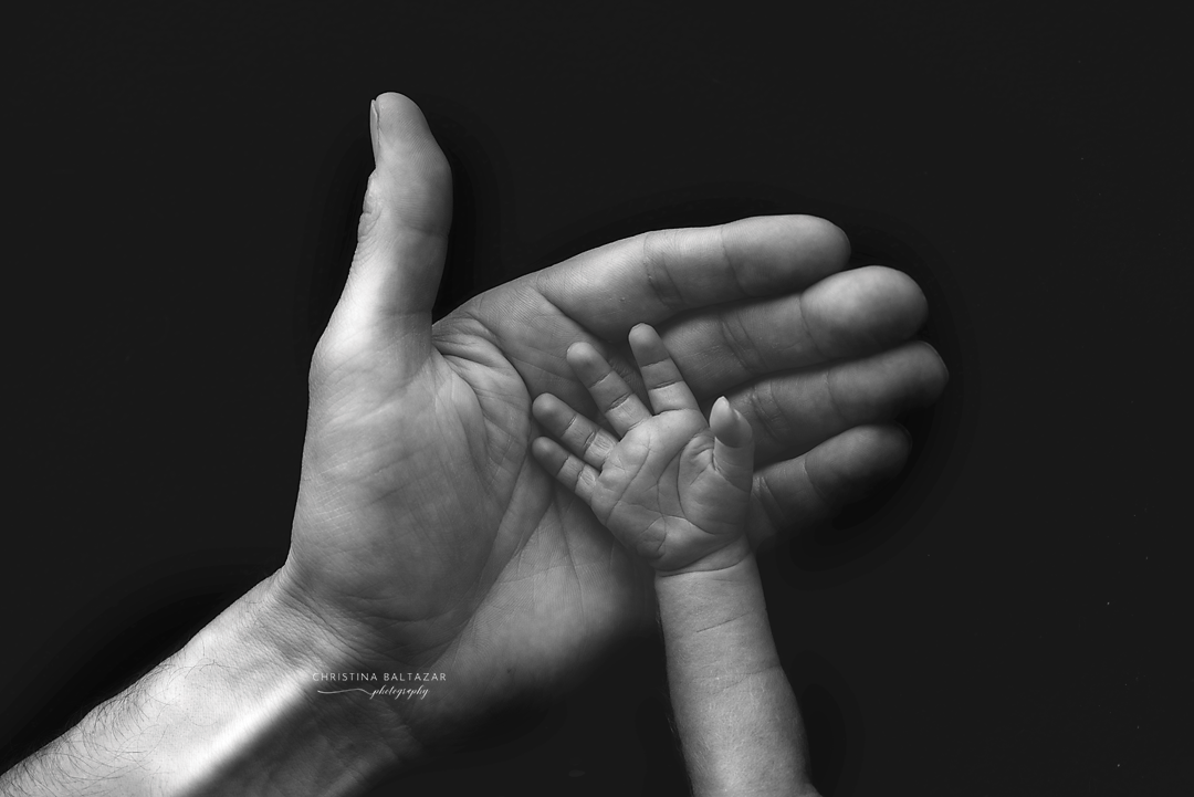Black and white photo of adult hand holding tiny baby hand against dark background.