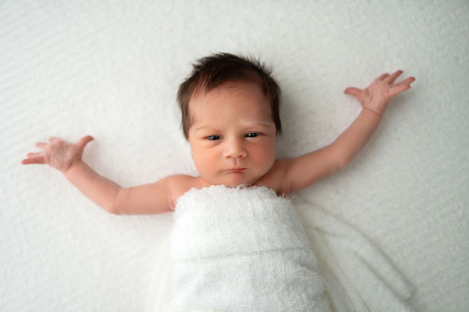 Newborn baby with arms outstretched wrapped in white blanket on light background.