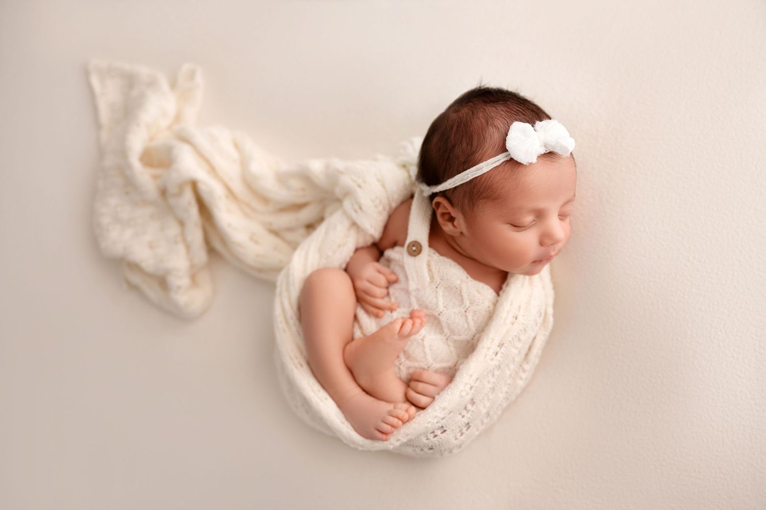 Newborn baby wrapped in soft white knit blanket sleeping peacefully with matching headband on cream background. Fresno Newborn Photography