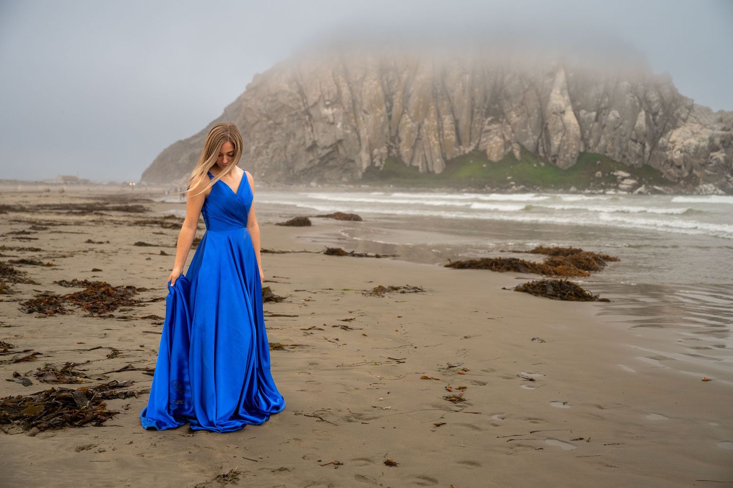 A flowing blue dress catches the wind on a misty beach with dramatic cliffs in the background.