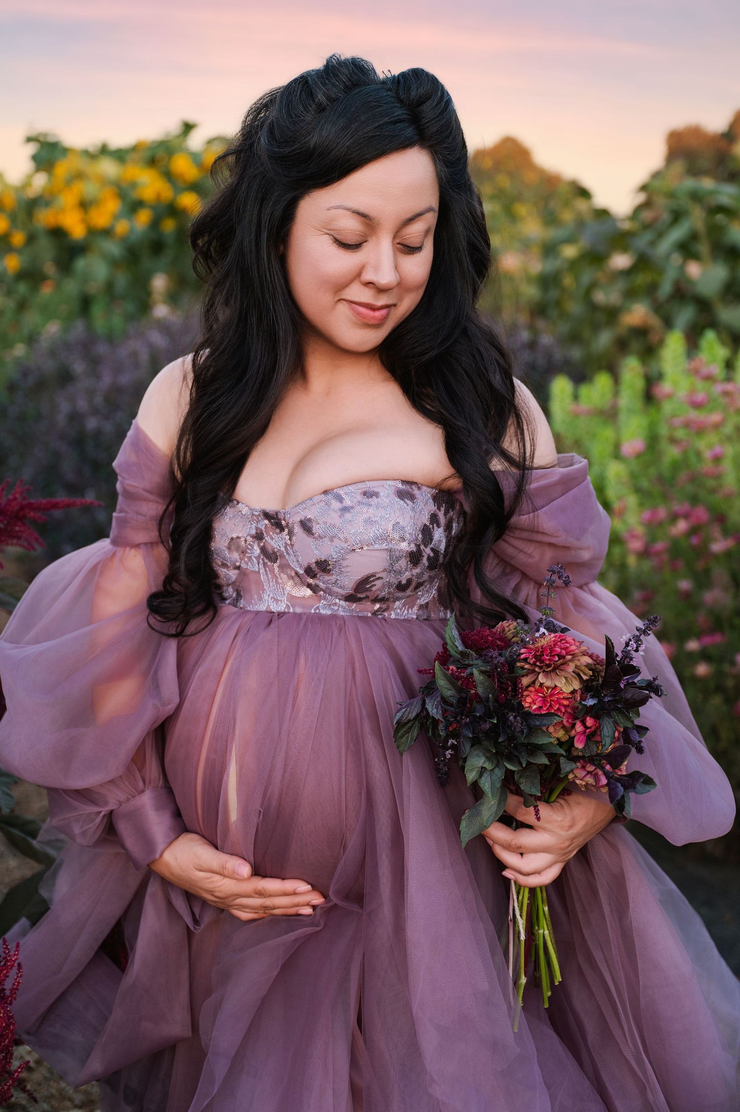 Maternity portraits in mauve tulle dress with floral bouquet in garden setting at sunset.