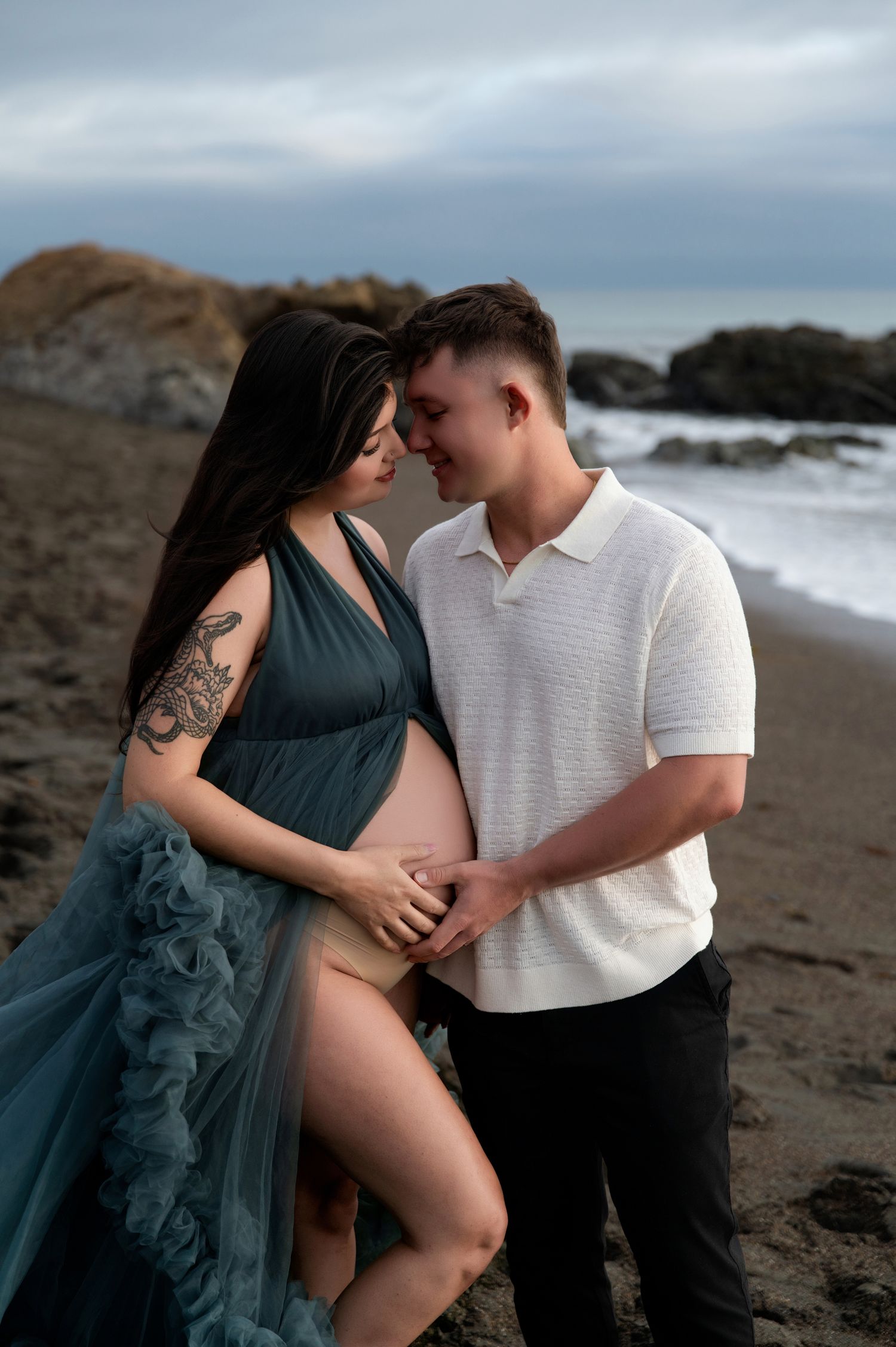 Romantic maternity photoshoot on a rocky beach with couple embracing in teal dress and white polo.