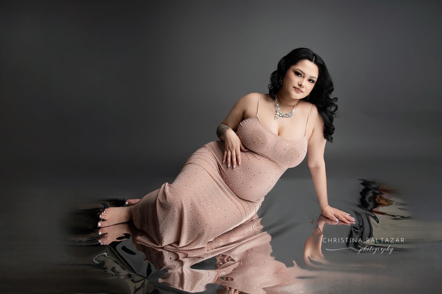 A figure in a flowing pink gown poses gracefully on the floor during an elegant maternity photoshoot. Hanford Maternity Photographer