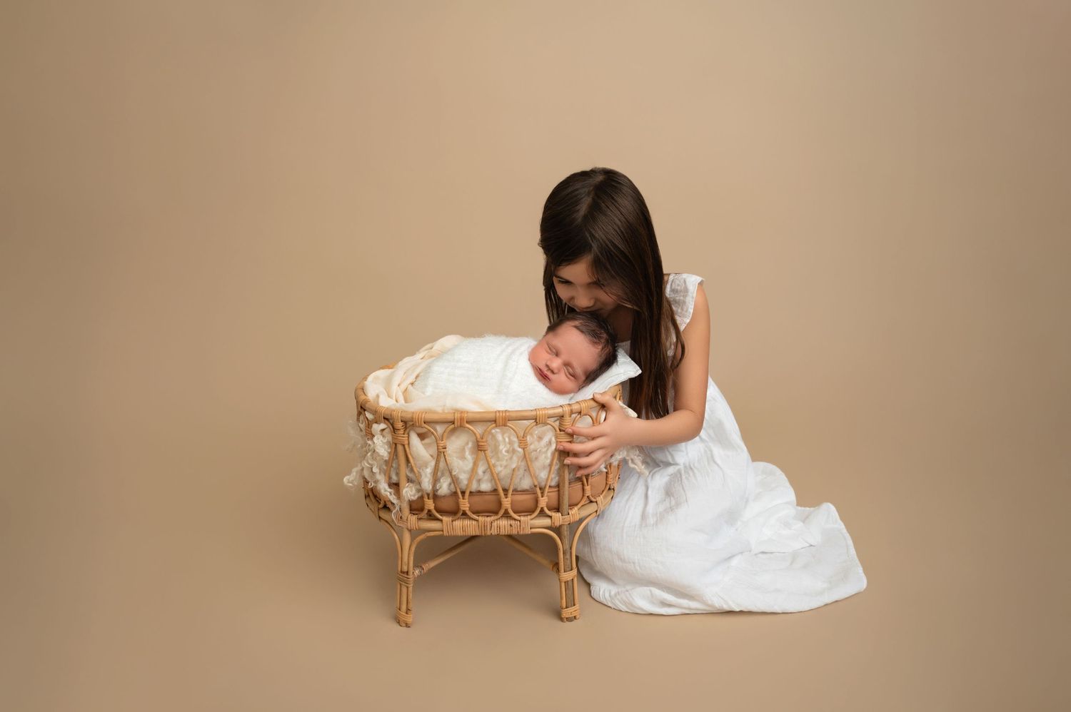 Girl in white dress gazing adoringly at baby sleeping in woven rattan bassinet against beige background.