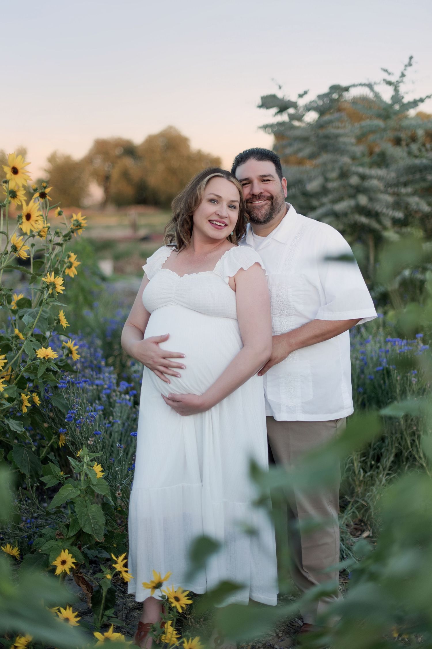 Pregnant couple smiling in a flower field with yellow sunflowers and purple blooms at sunset, woman in white dress.