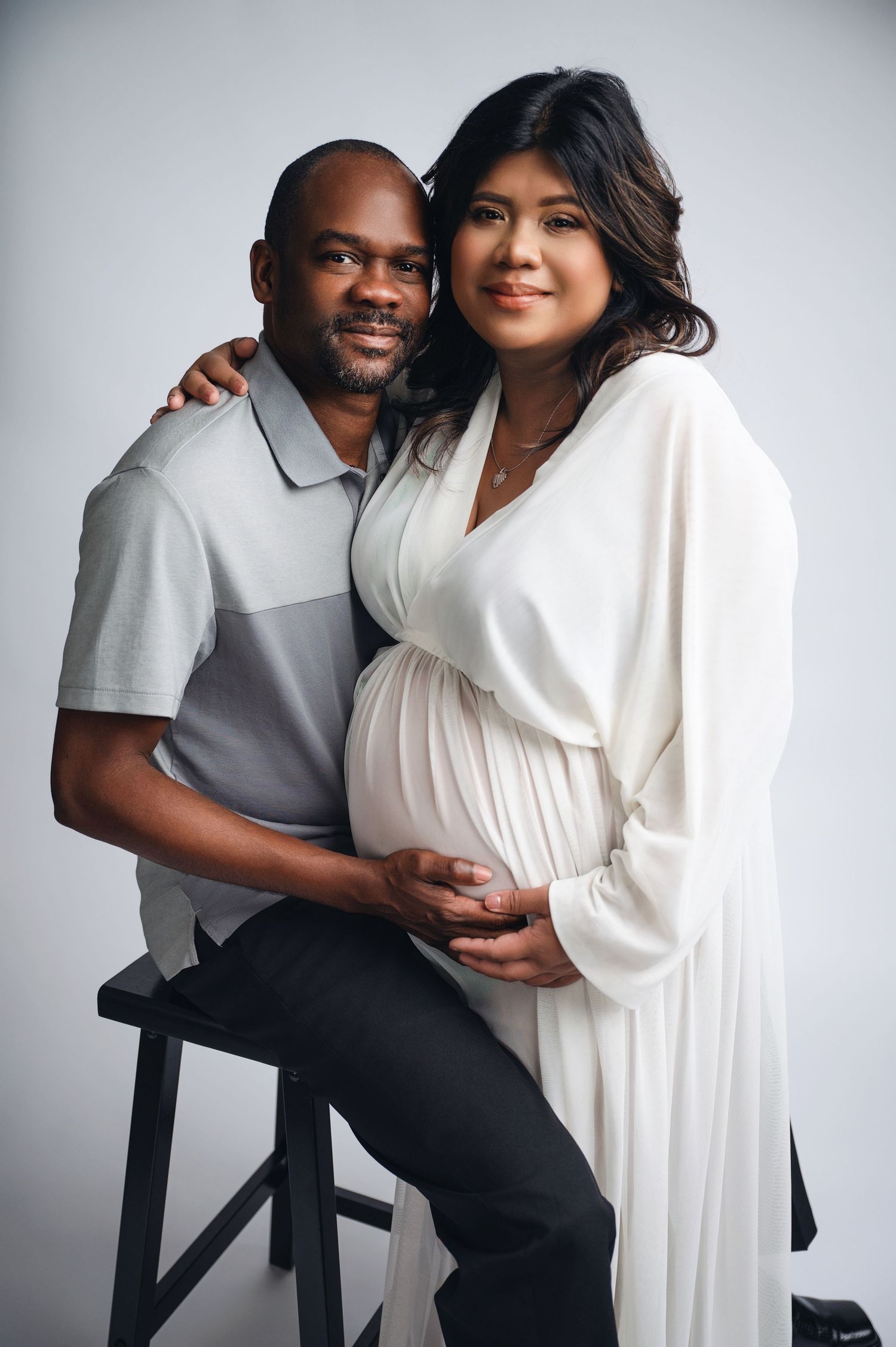 Couple in white and grey casual wear posing against white studio background. Hanford Maternity Photographer
